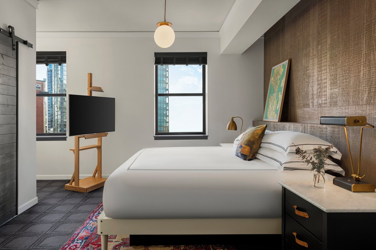 A modern bedroom with a large bed, a desk with a lamp, a map on the wall, and a view of buildings through the window.