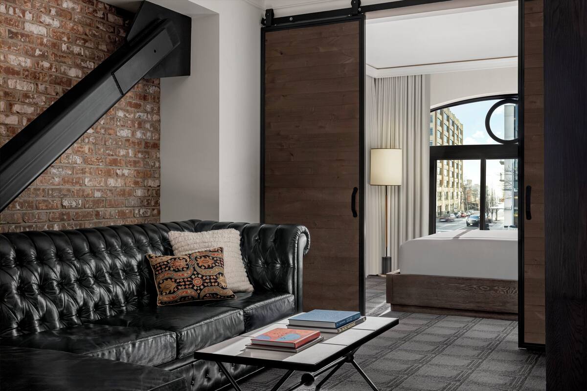 A modern living room with a black leather sofa, a coffee table with books, a brick wall, and a view of the city through a large window.
