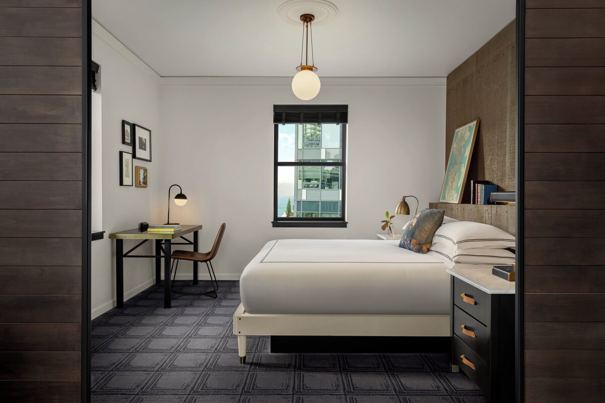 A bedroom with a bed, bedside table, lamp, chair, and a window with a view.