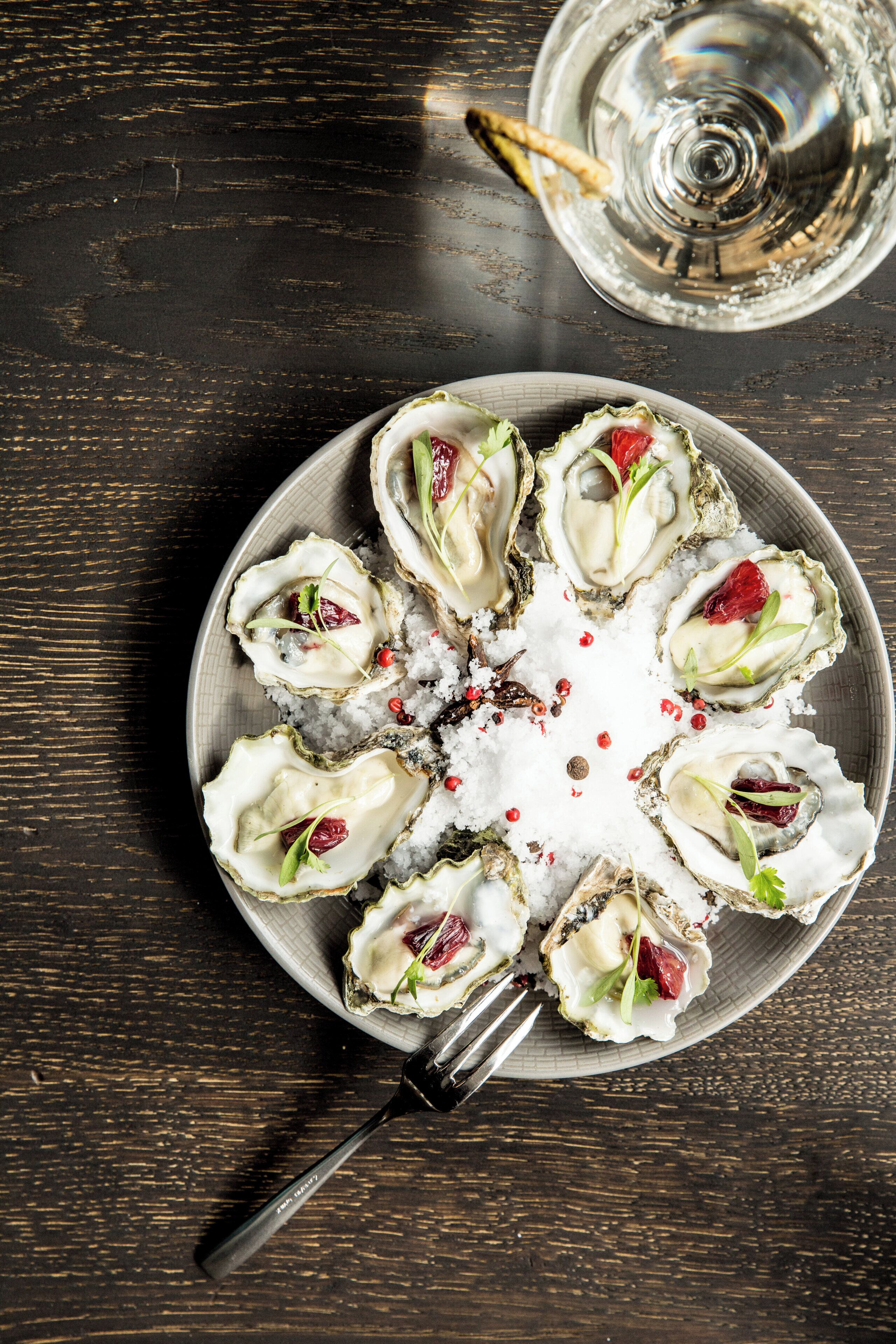 A plate of oysters garnished with herbs and red berries, served on a bed of salt, with a glass of water and a fork on a wooden table.