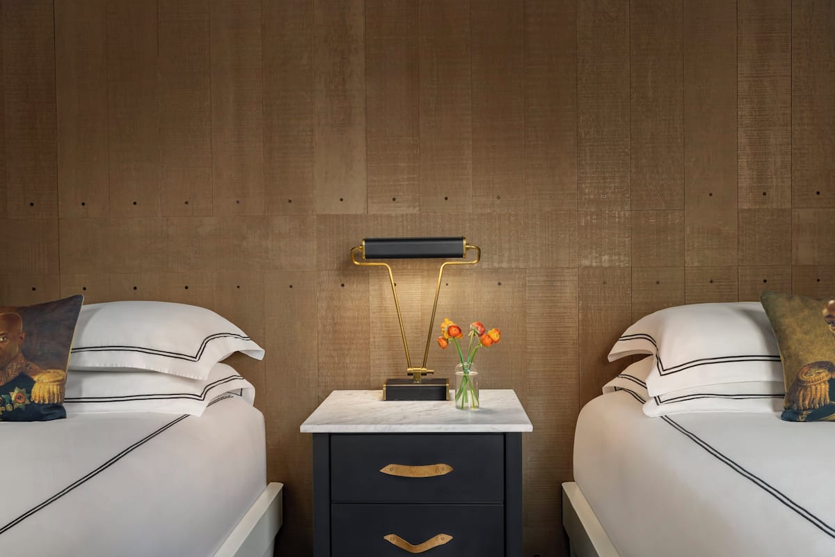 A hotel room with two beds, a nightstand with a vase of flowers, and a wall-mounted lamp.