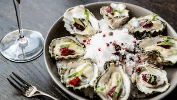 Un plato de ostras adornado con hierbas y especias, acompañado de una copa de vino blanco sobre una mesa de madera oscura.