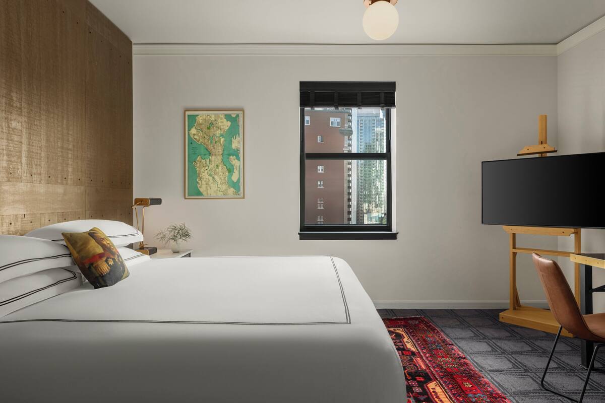 A bedroom with a bed, a map on the wall, a window with a view of buildings, and a desk with a chair.