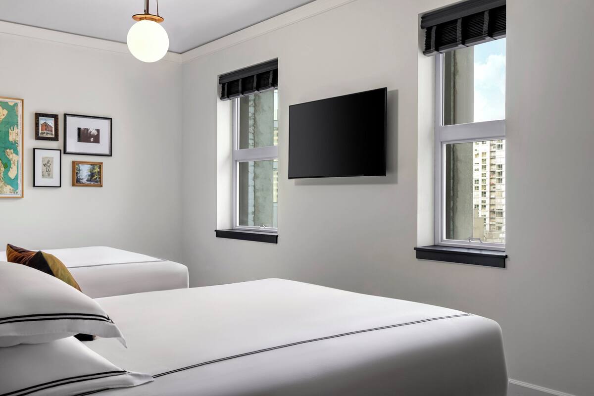 A modern hotel room with two beds, a flat-screen TV, and large windows with blinds.