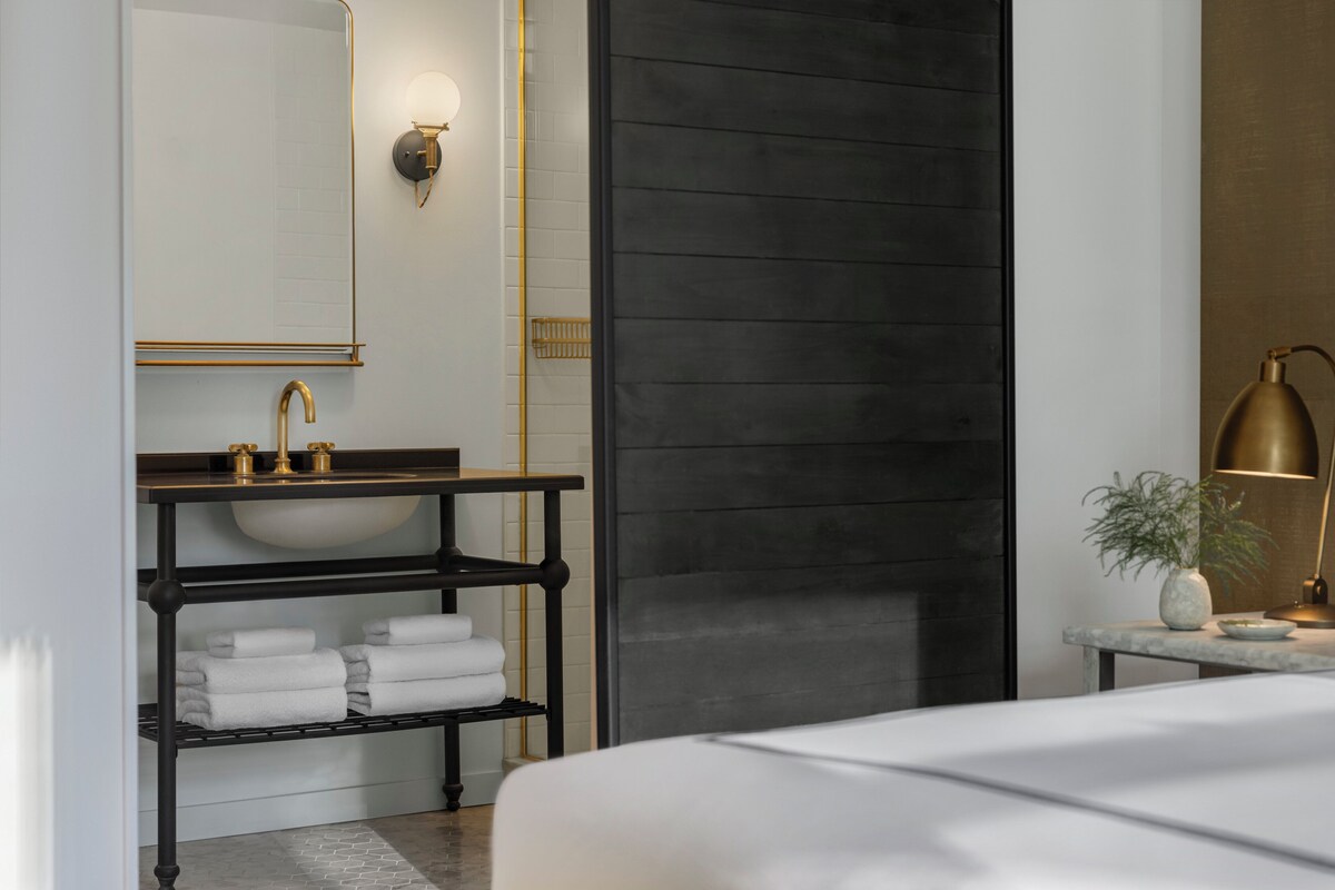A modern bathroom with a large mirror, a gold faucet sink, a black shelf with towels, and a marble countertop with a lamp.