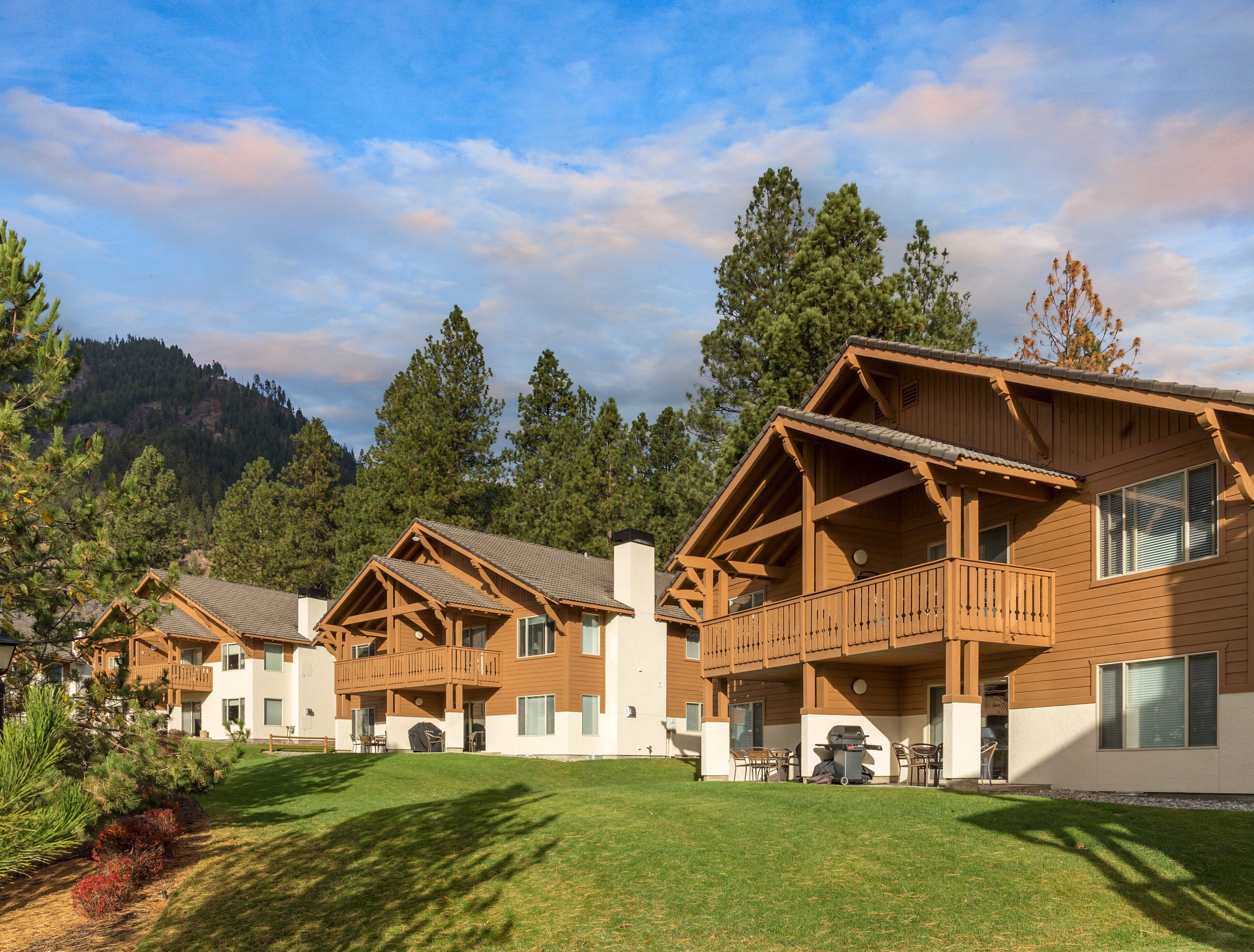 Una hilera de cabañas de madera con balcones, rodeadas de un césped cuidado y un bosque denso.