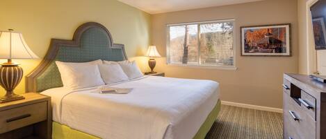 A neatly made bed with white linens, a green upholstered headboard, and a nightstand with a lamp. There is a window with blinds, a framed picture on the wall, and a dresser with drawers.