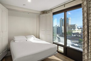 A bedroom with a large bed, white walls, and a view of a cityscape through a large window.