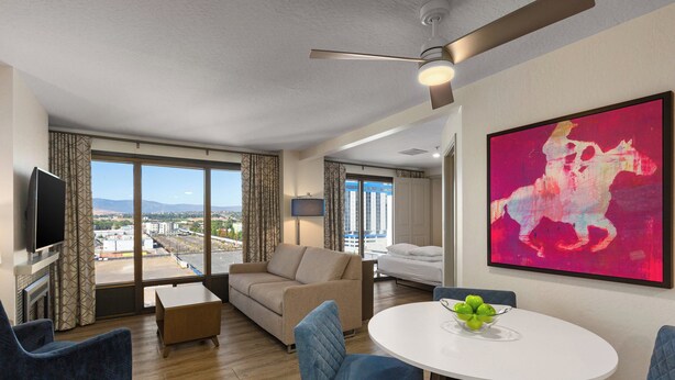 Een moderne hotelkamer met een groot raam, een flatscreen televisie, een ronde eettafel met blauwe stoelen, een bank en een plafondventilator.
