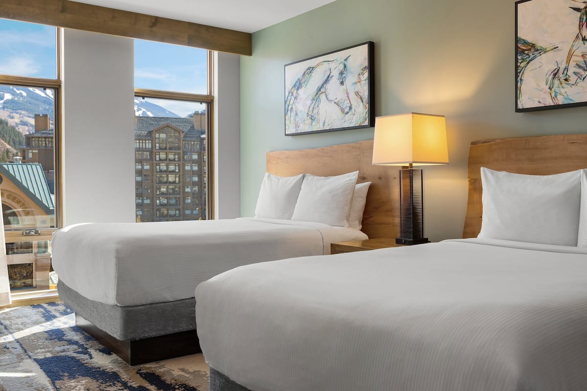 A hotel room with two beds, a large window with a mountain view, and artwork on the walls.