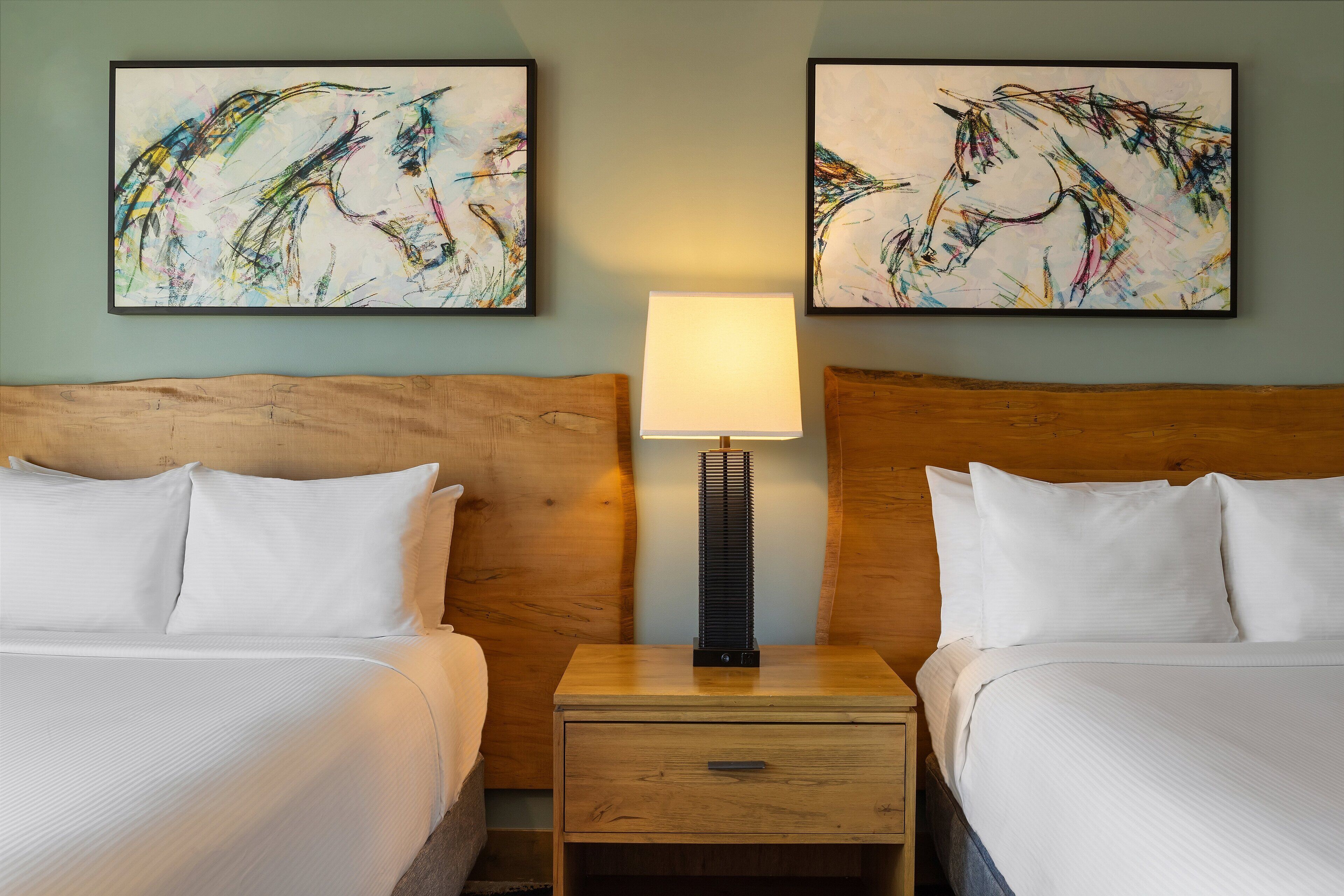Een hotelkamer met twee bedden, een nachtkastje met een lamp en twee abstracte schilderijen aan de muur.