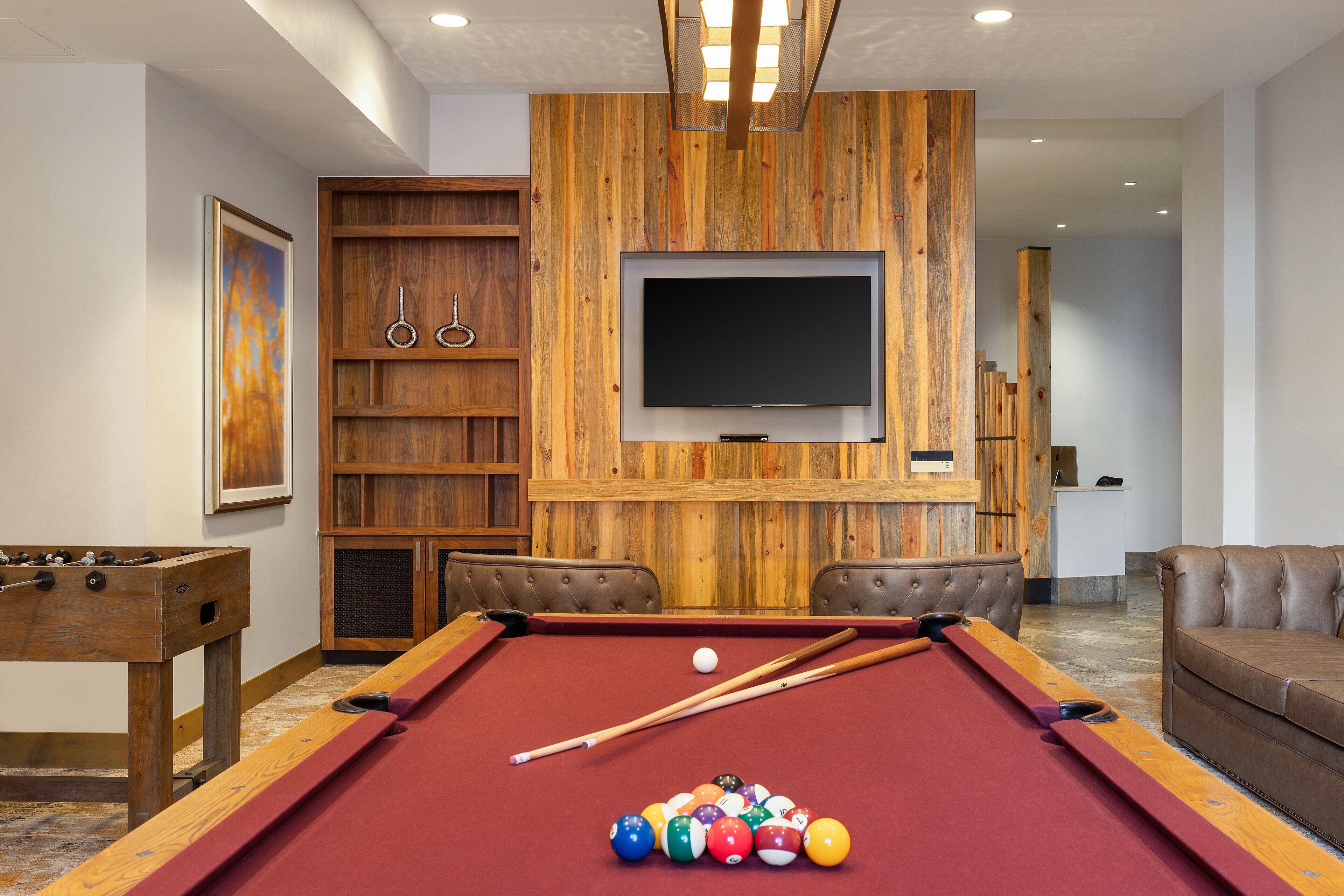 A pool table with balls, a foosball table, and a television mounted on a wooden wall.
