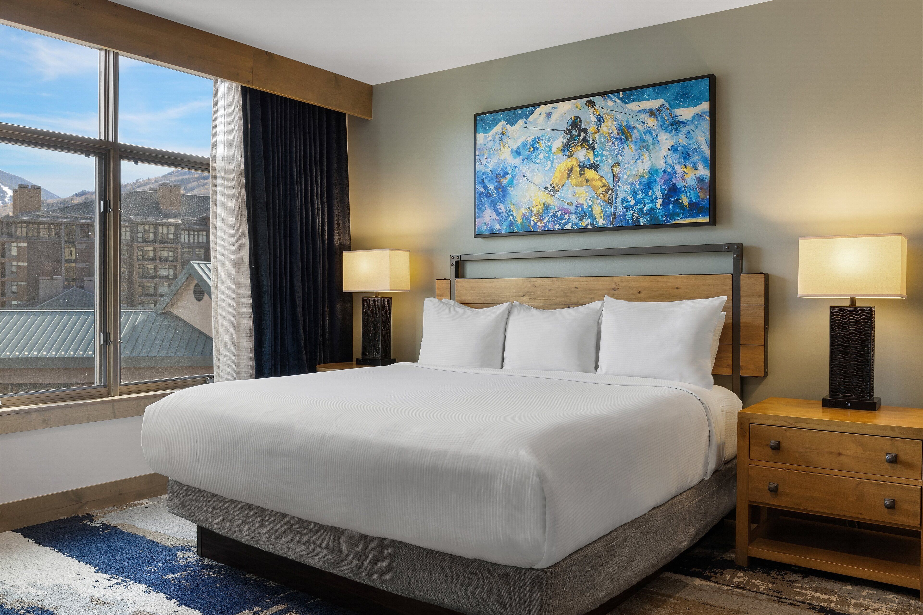 A hotel room with a large bed, two bedside tables with lamps, a window with a view of buildings, and a painting on the wall.