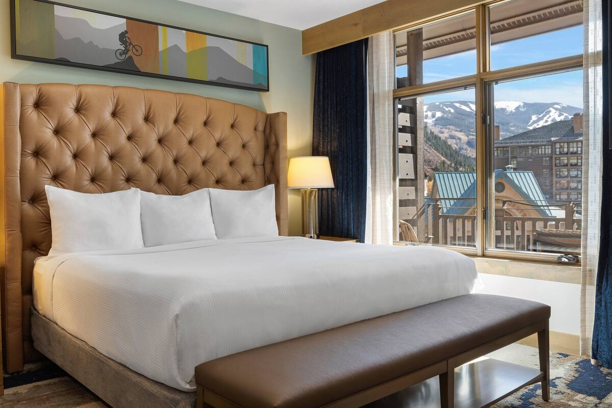 A large bed with a tufted headboard, a bedside lamp, and a view of mountains through a window.