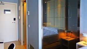 Habitación de hotel con cama, mesita de noche con lámpara, una silla y una puerta.