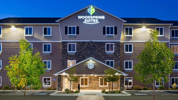 Un hôtel à plusieurs étages, reconnaissable à son grand panneau « Woodspring Suites ». Il se distingue par son entrée centrale, sa façade en pierre et ses nombreuses fenêtres.