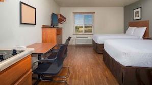 Une chambre d’hôtel avec deux lits, un bureau avec une chaise, une petite kitchenette et une fenêtre avec des stores.