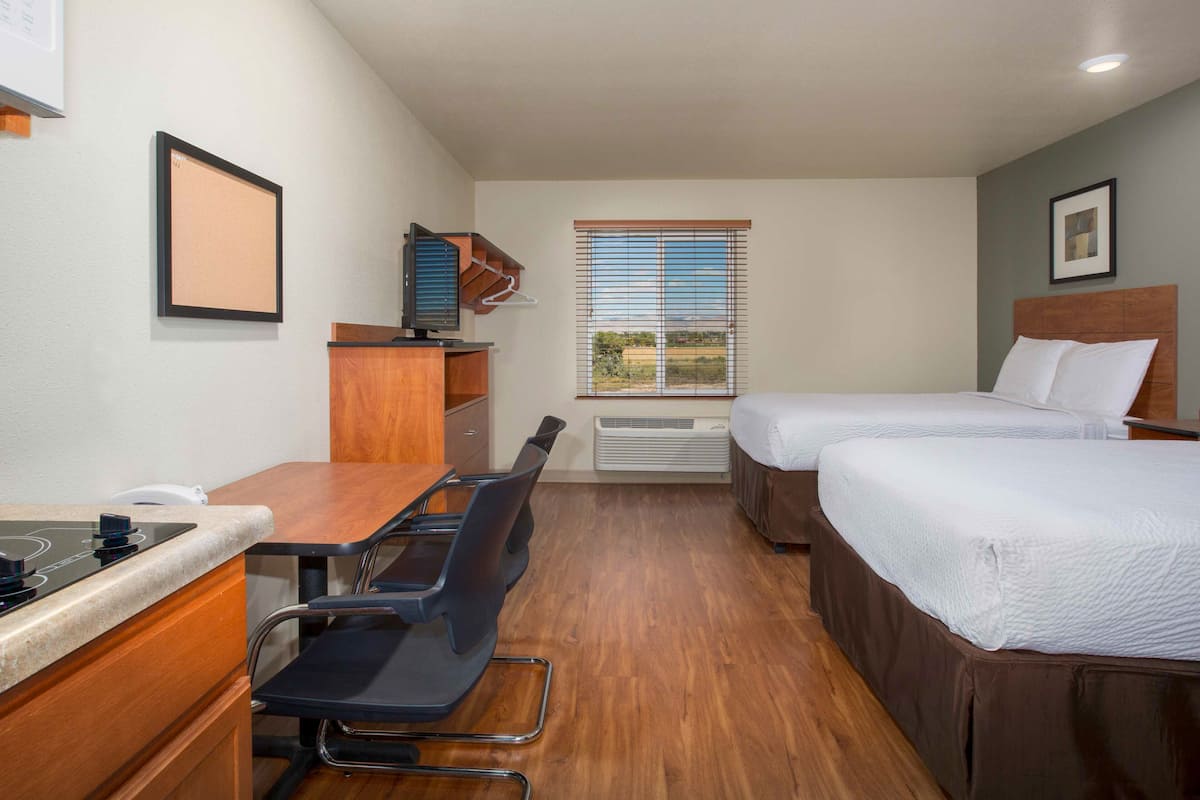 Ein Hotelzimmer mit zwei Betten, einem Schreibtisch mit Stuhl, einer kleinen Küchenzeile und einem Fenster mit Jalousien.