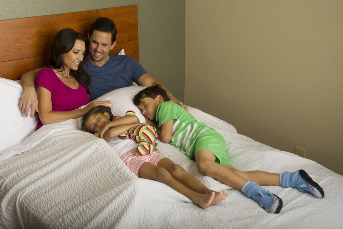 Eine Familie mit drei Personen auf einem Bett mit einem hölzernen Kopfteil, einer weißen, strukturierten Decke und einer schlichten Wand im Hintergrund.