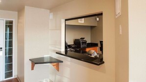 Un couloir avec un plancher en bois, une petite étagère noire et un miroir qui reflète une imprimante et un bureau.