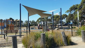 Un parque infantil con zona sombreada, juegos cercados y suelo de arena.