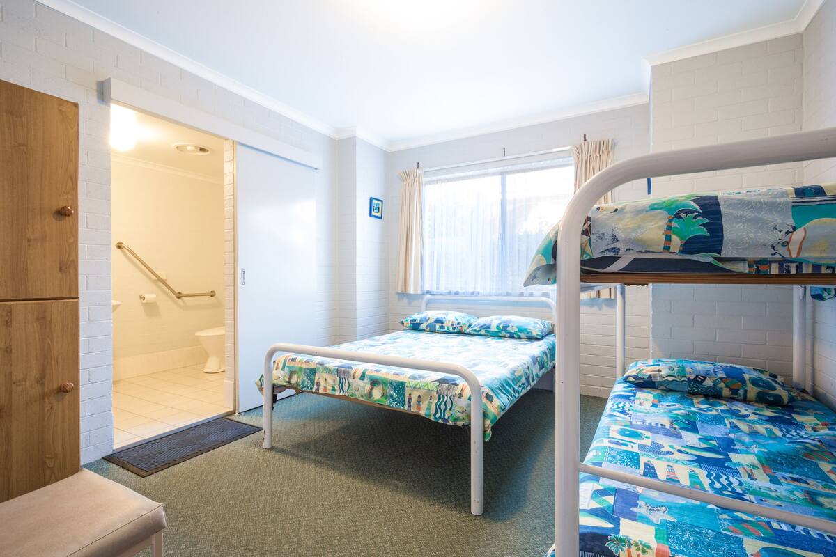 A room with a bunk bed, a single bed, a wooden wardrobe, a door leading to a bathroom, and a window with curtains.