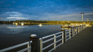 Un muelle frente al agua con pasarela de madera y barandillas blancas, barcos amarrados al final, y una masa de agua tranquila que refleja el cielo.