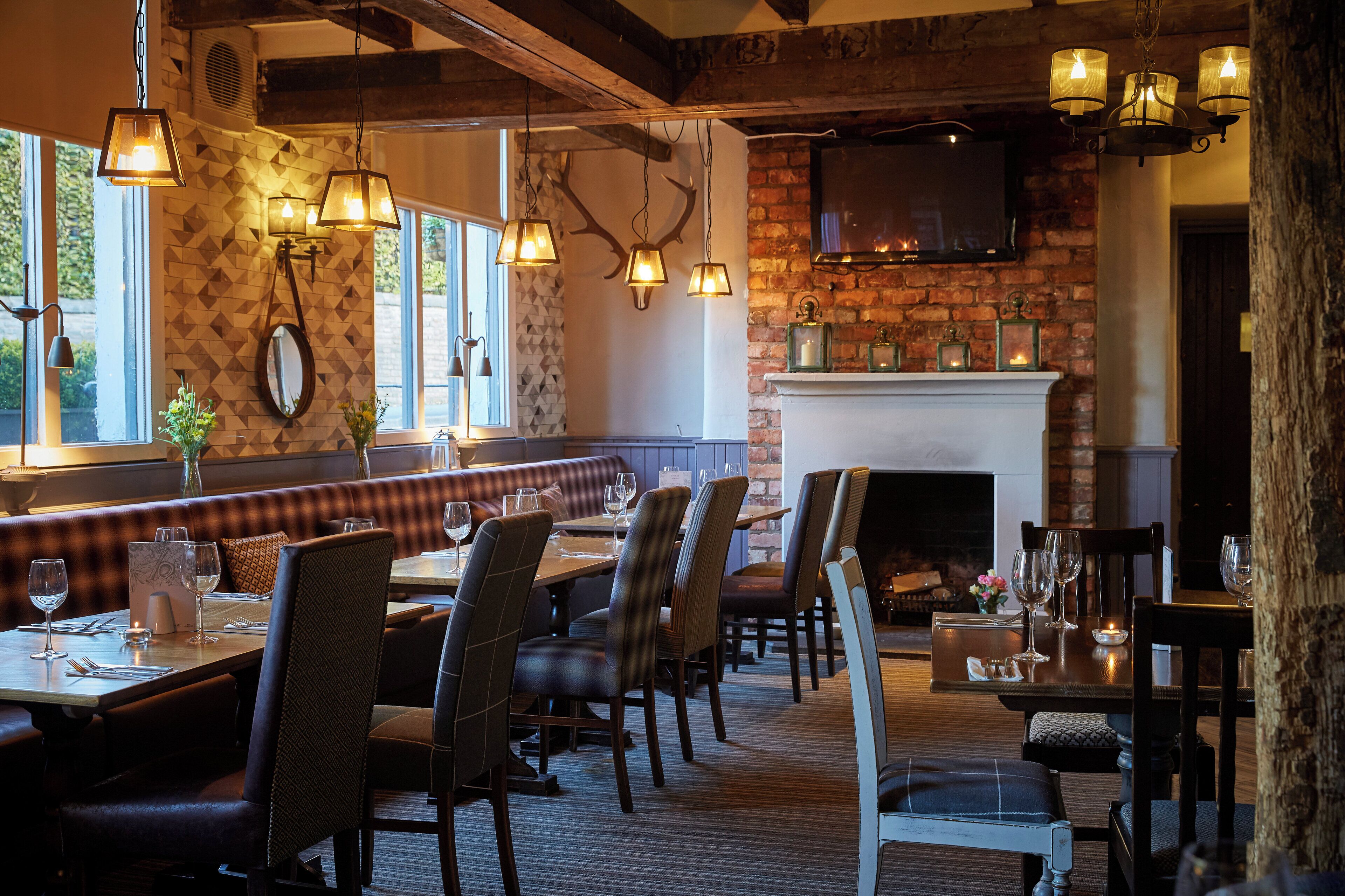 L’intérieur d’un restaurant présente des murs en briques, des poutres en bois et une cheminée. Les tables sont dressées avec des verres à vin et des menus.