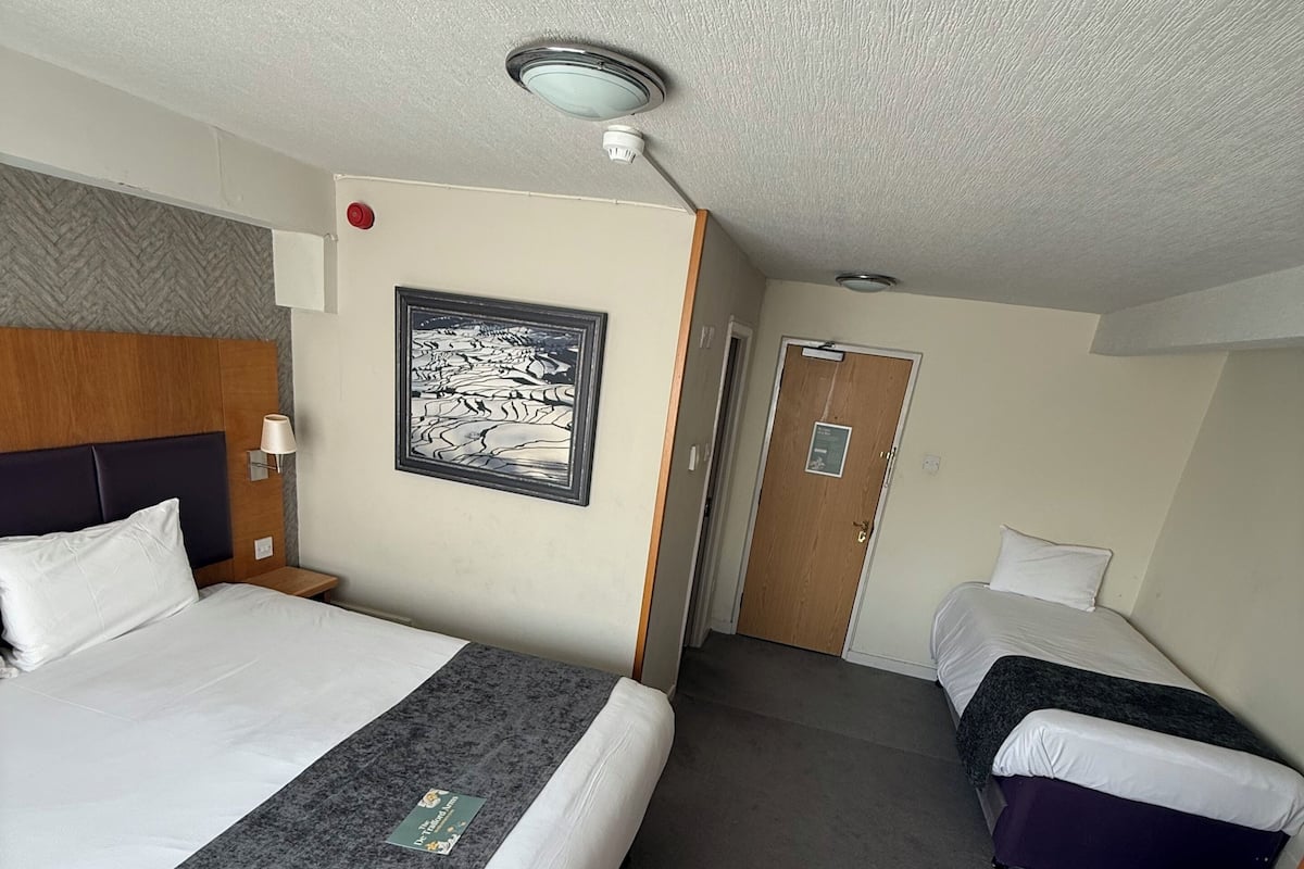 Habitación de hotel con una cama grande, una cama más pequeña, un escritorio con papeles, una lámpara de pared y un cuadro enmarcado.