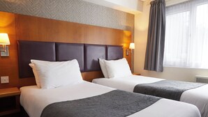 Hotellihuone, jossa on kaksi sänkyä, puiset päätyosat, valkoiset vuodevaatteet ja ikkuna, jossa on verhot.
