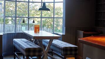 A corner seating area with a table and benches, a hanging light fixture, and a view through large windows.