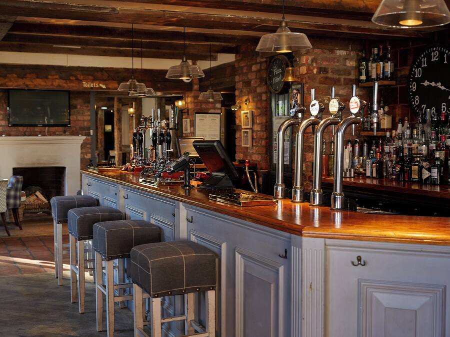 A bar area with a wooden counter, multiple taps, stools, and a brick wall.