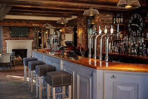 Uma área de bar com balcão de madeira, várias torneiras, banquetas e parede de tijolos.