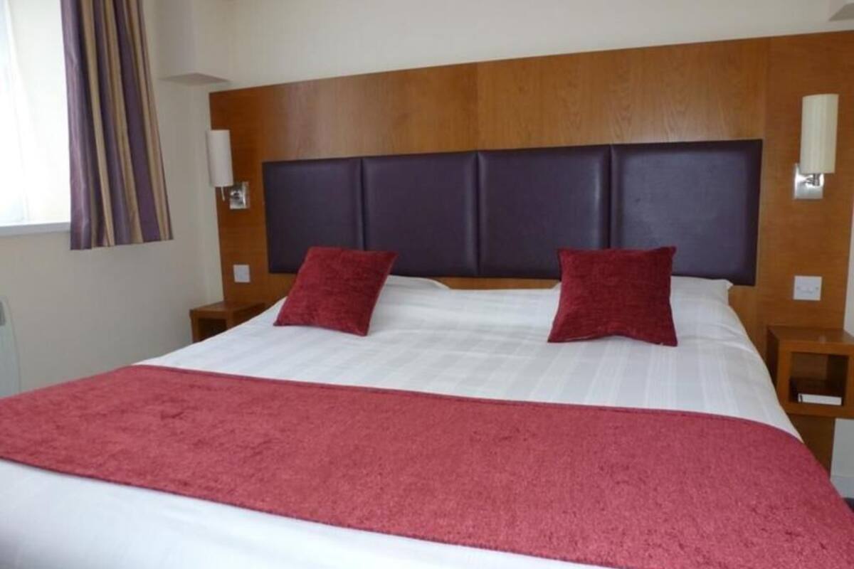 Una habitación de hotel con una cama grande, un cabecero de madera, dos mesitas de noche y una manta roja.