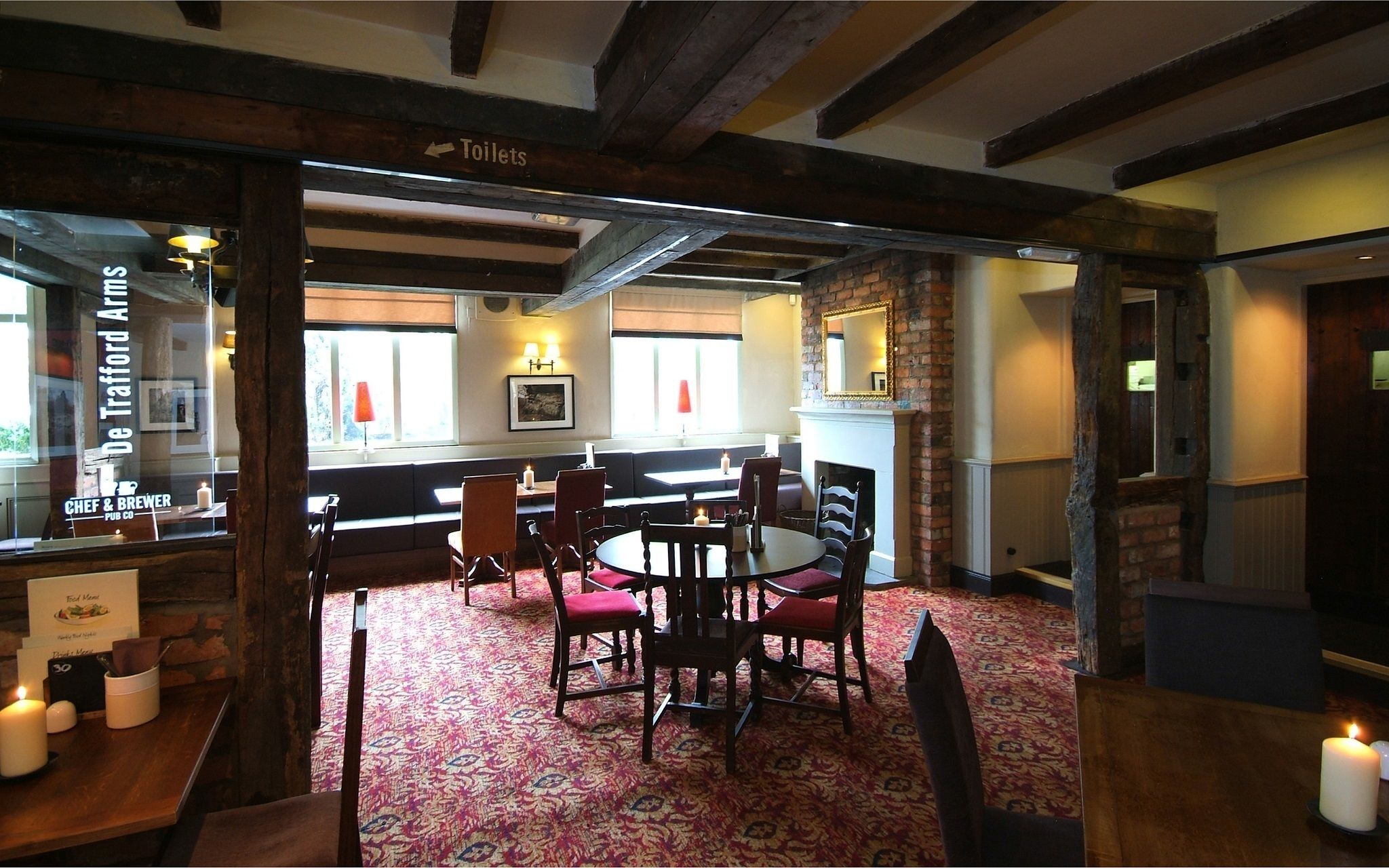 A cozy pub interior with a round table and chairs, a brick fireplace, and a view of the outside through large windows.