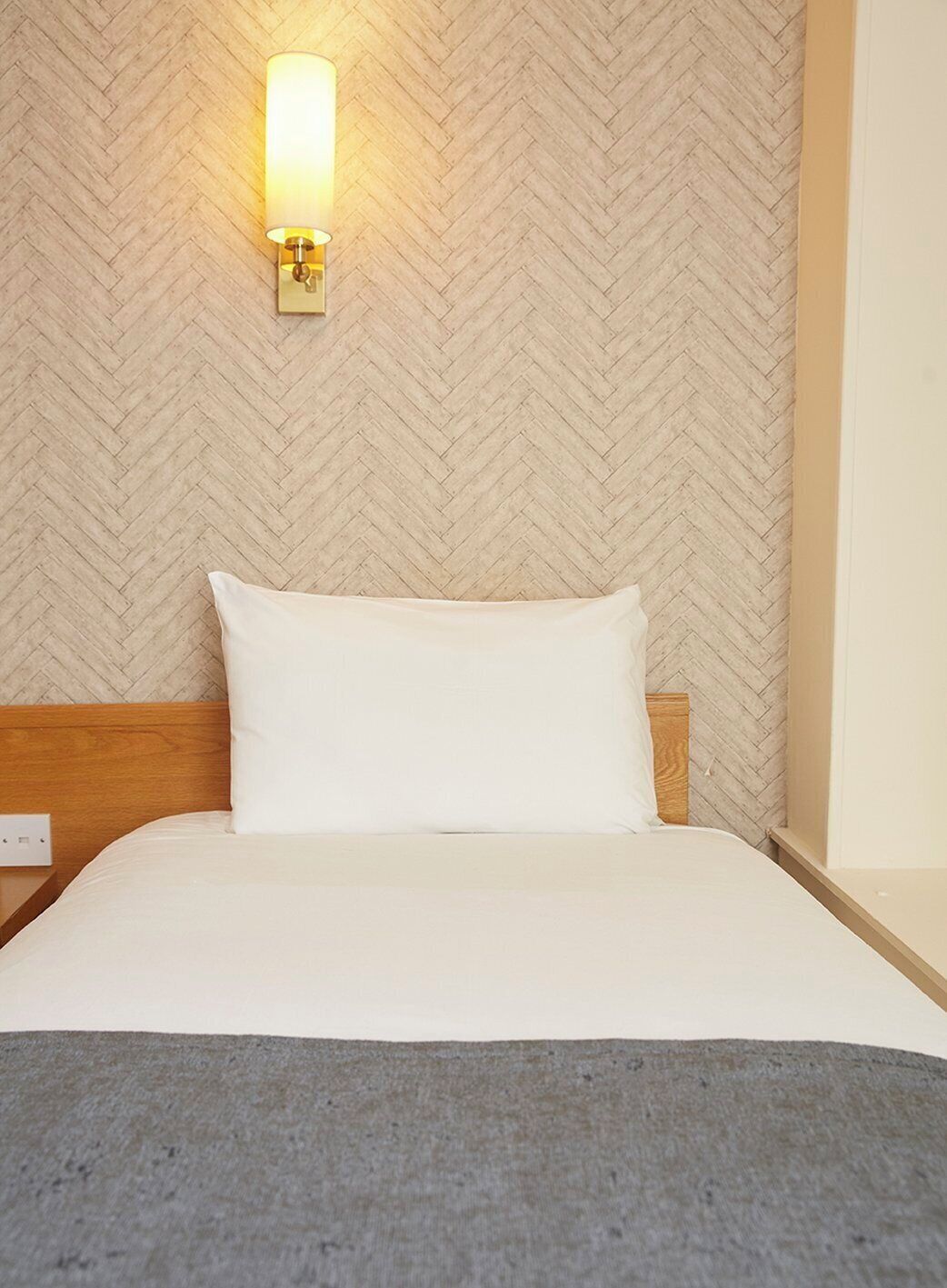 Una cama bien hecha con un almohadón blanco y una manta gris, cabecera de madera y una lámpara de pared con diseño en espiga.