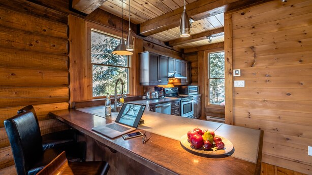 A wooden cabin interior with a kitchen area, a dining table with chairs, and a fruit bowl on the counter. - RCNT Chalets (Lac-Superieur)