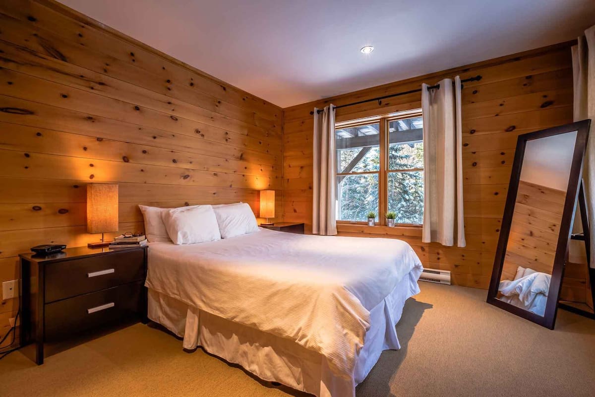 A wooden-paneled bedroom with a bed, bedside lamps, a nightstand, and a window with curtains.