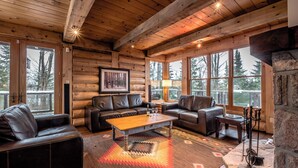 A log cabin interior with leather sofas, a wooden coffee table, and large windows offering views of the forest. - RCNT Chalets (Lac-Superieur)