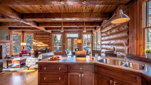 A log cabin interior with a wooden dining area, a kitchen with a large island, and a stone fireplace. - RCNT Chalets (Lac-Superieur)
