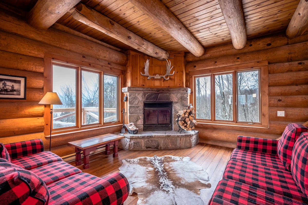 A log cabin interior with a stone fireplace, plaid furniture, and a large window overlooking a snowy landscape.