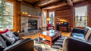 A log cabin interior with a stone fireplace, leather furniture, and large windows offering views of trees. - RCNT Chalets (Lac-Superieur)