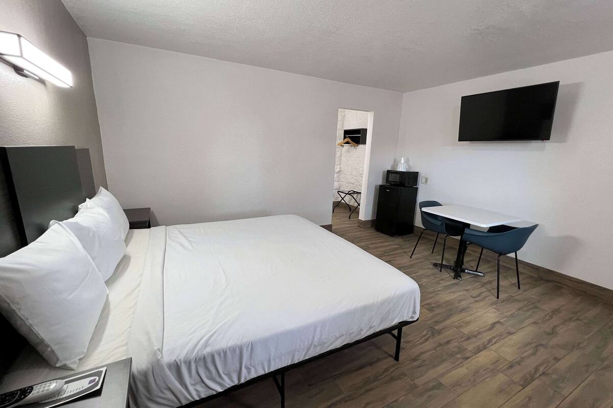 A hotel room with a bed, a desk with a chair, a TV mounted on the wall, and a small kitchenette area.