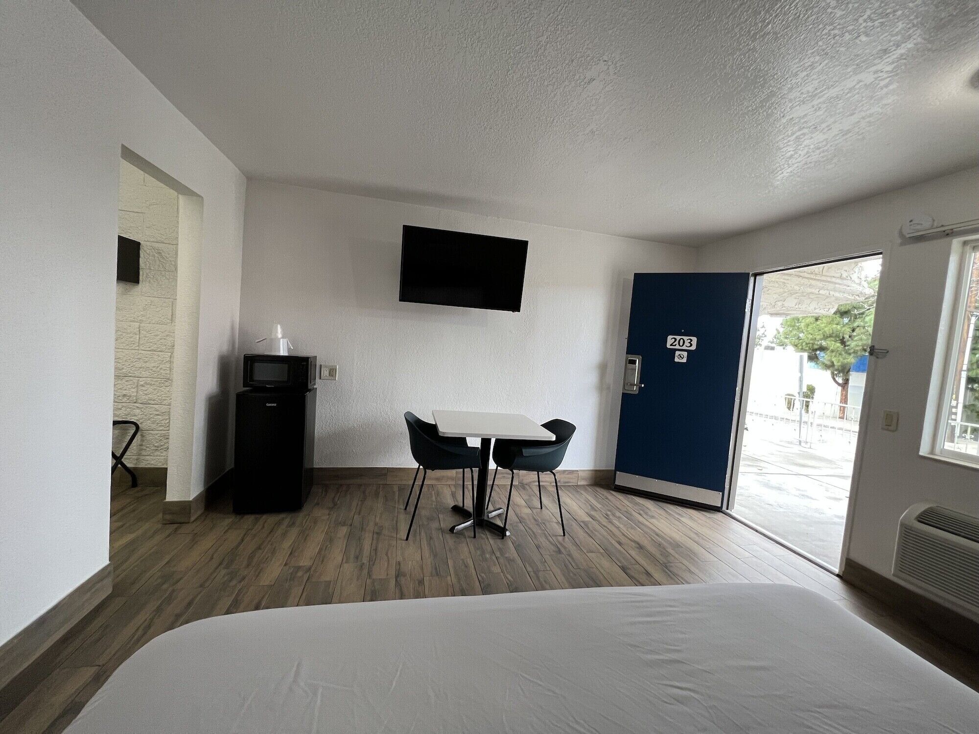 A hotel room with a bed, a desk with a chair, a microwave, a TV mounted on the wall, and a door with the number 203.