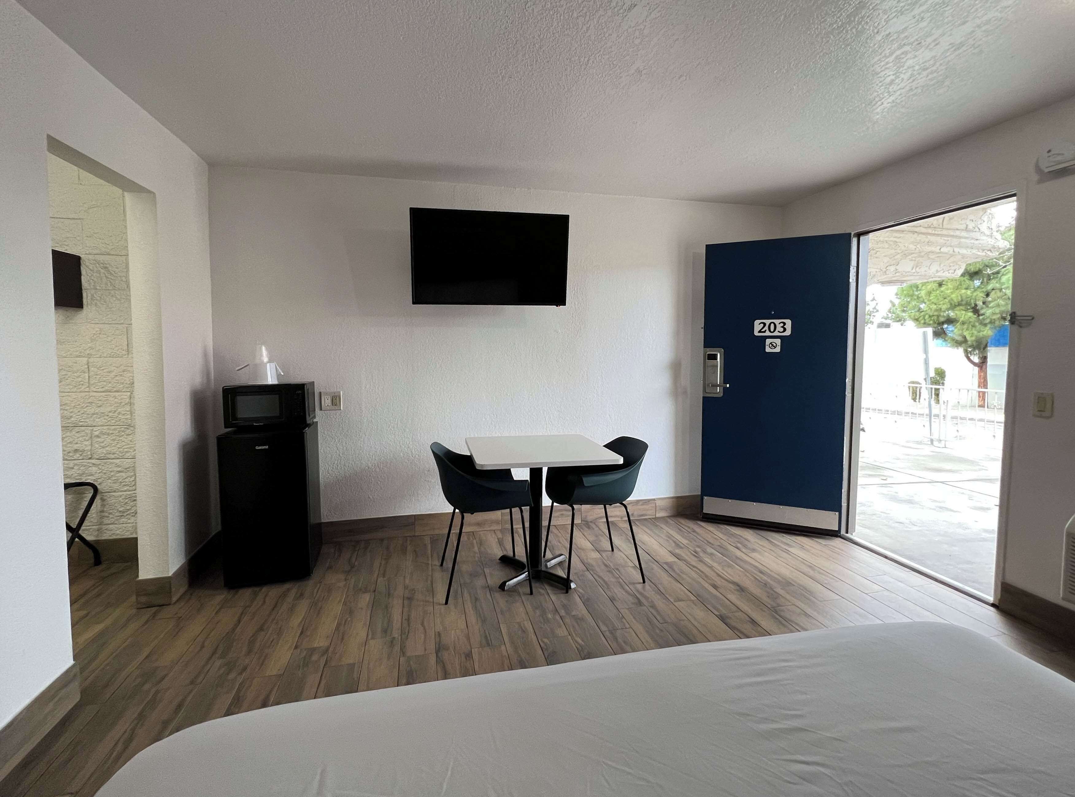 A hotel room with a bed, a TV mounted on the wall, a small desk with a chair, and a door with a number.