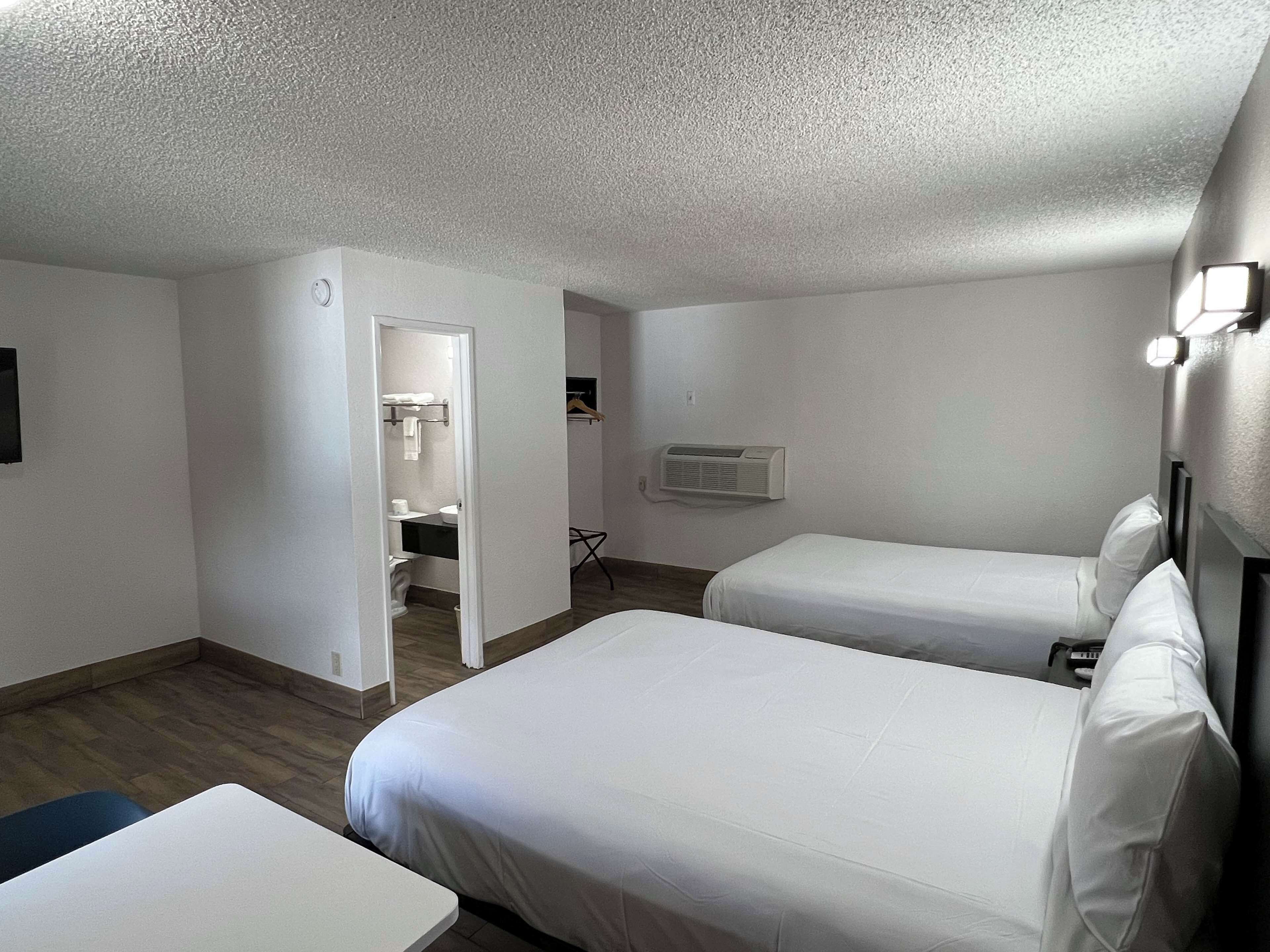 A hotel room with two single beds, a small table, a bathroom with a toilet and sink, and an air conditioning unit.