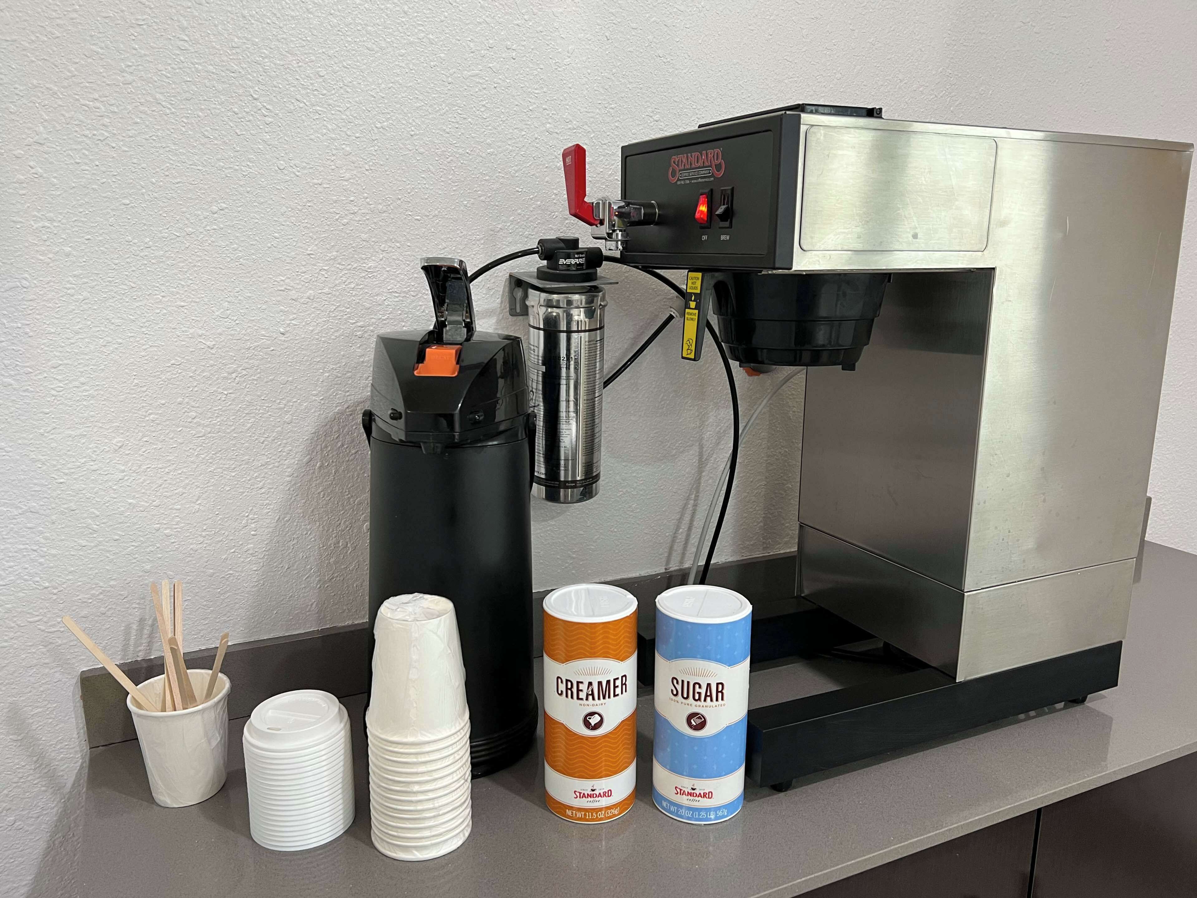 Una cafetera con vasos desechables y dos recipientes para líquidos, uno etiquetado 'CREAMER' y otro 'SUGAR'.