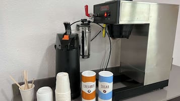 Una cafetera con vasos desechables y dos recipientes para líquidos, uno etiquetado 'CREAMER' y otro 'SUGAR'.
