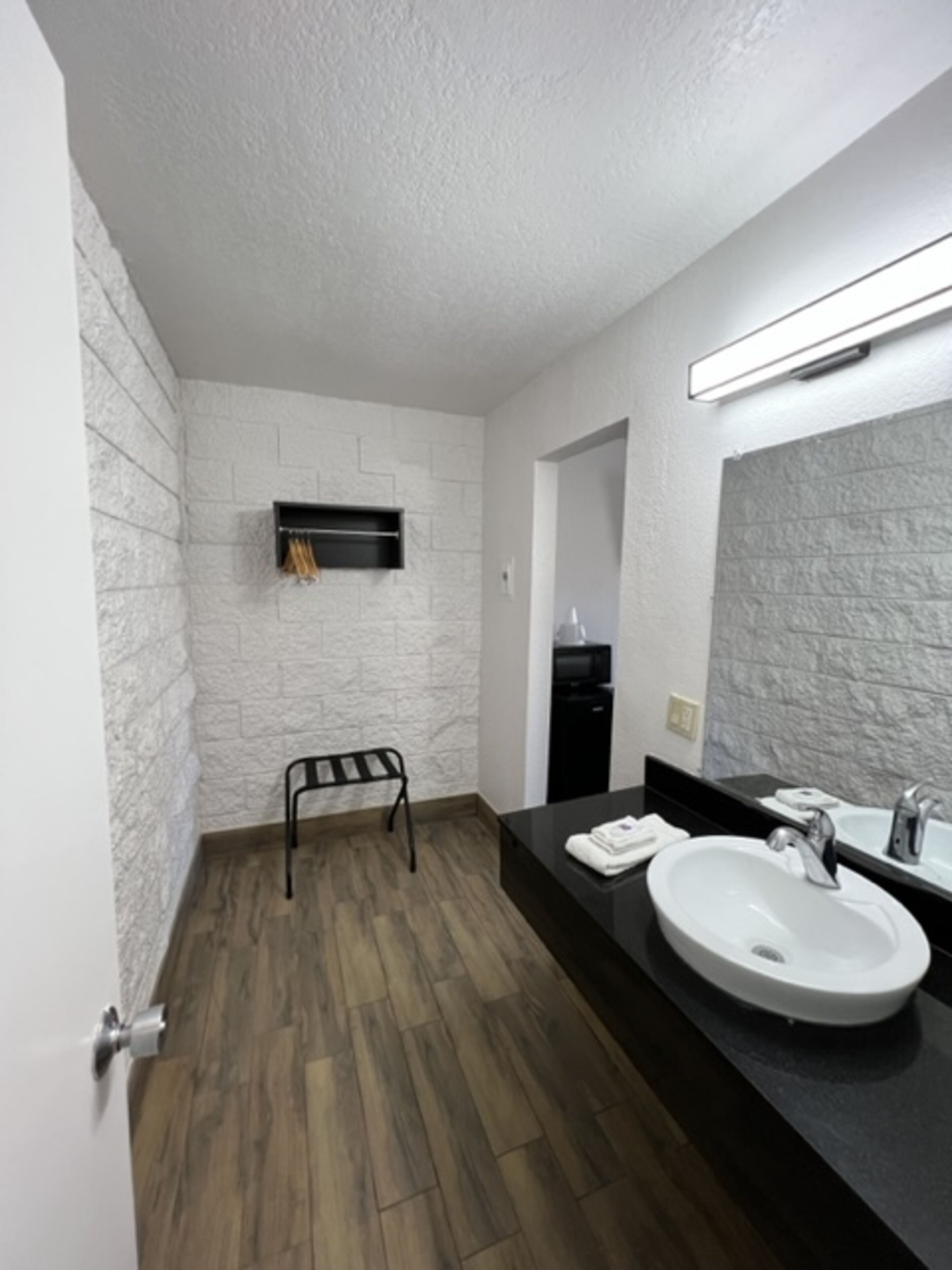 Un baño moderno con una pared blanca con textura, suelo de madera, toallero negro, encimera negra con lavabo blanco y un espejo grande.