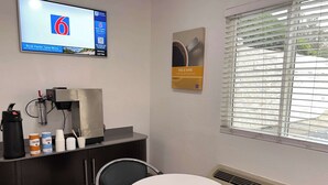 A small, well-lit room with a coffee machine, a table, chairs, and a TV displaying a booking interface. - Motel 6 Orange, CA - Anaheim (Orange)